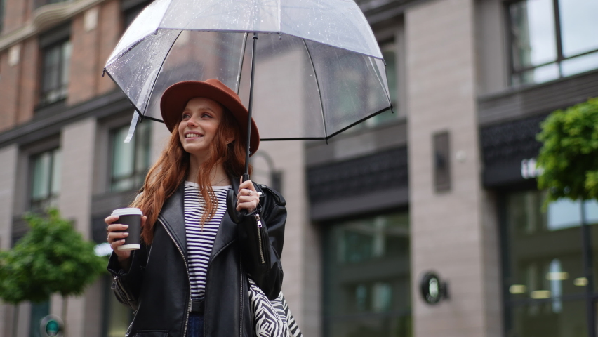 Low-angle view of attractive satisfied young woman in hat standing with transparent umbrella in city cold rain and drinking hot coffee. Female tourist enjoying drink in rainy weather by building.