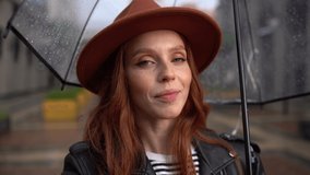 POV of cheerful young woman having video call using smartphone holding transparent umbrella standing in city in autumn rain. Close-up face of cute lady talking online videocall looking at camera.