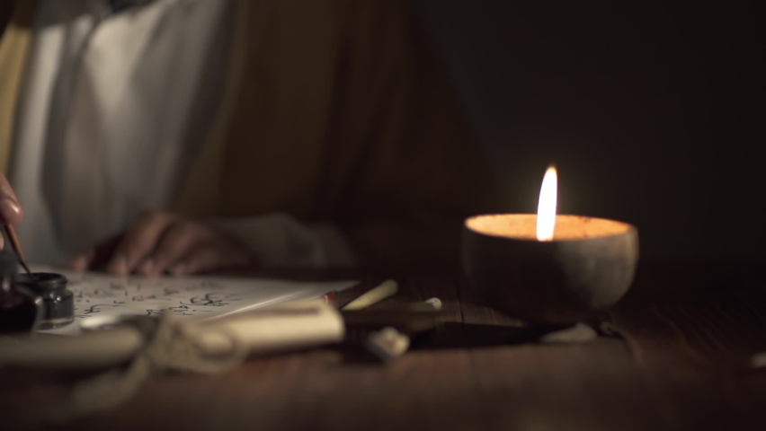 A medieval man uses an old pen with ink to write Arabic script on an a paper lit by a candle,slow motion