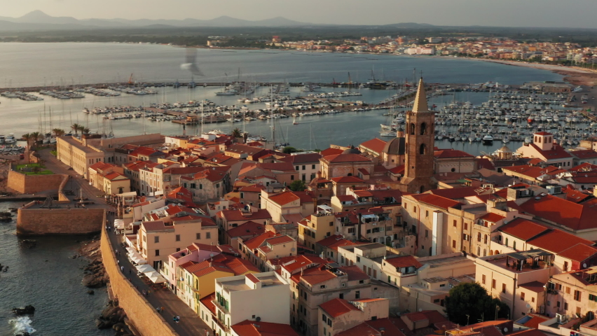 Cinematic shot of the old town of Alghero, Sardinia - Italy.
Drone view of ancient Italian city at Sunset. Golden hour, warm colors, view of the harbor in the distance.