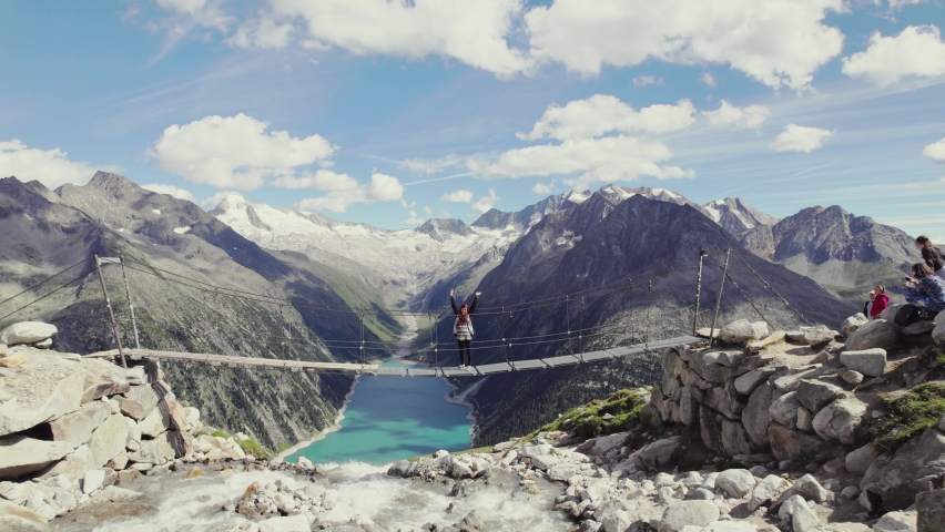 Beautiful redhead girl standing on a wooden bridge waving. massive alpine mountain range scenery with glaciers in the distance. Big Schlegeis reservoir barrier lake in the valley on a sunny day 4K.