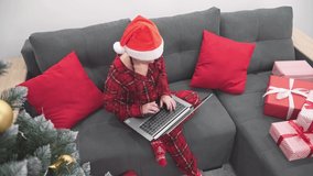 A girl in Santa Claus hat communicates via video call on computer. Celebrating home online. Woman in red on sofa at home with laptop, Christmas tree and gifts. New Year holidays shopping and ordering.