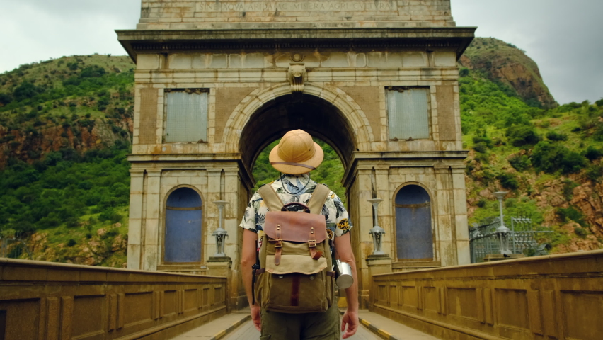 a male traveler with a backpack and a cap stands on a bridge near the historical arch. male tourist in safari clothes near triumphal arch. a man walks across a bridge with a backpack and shorts