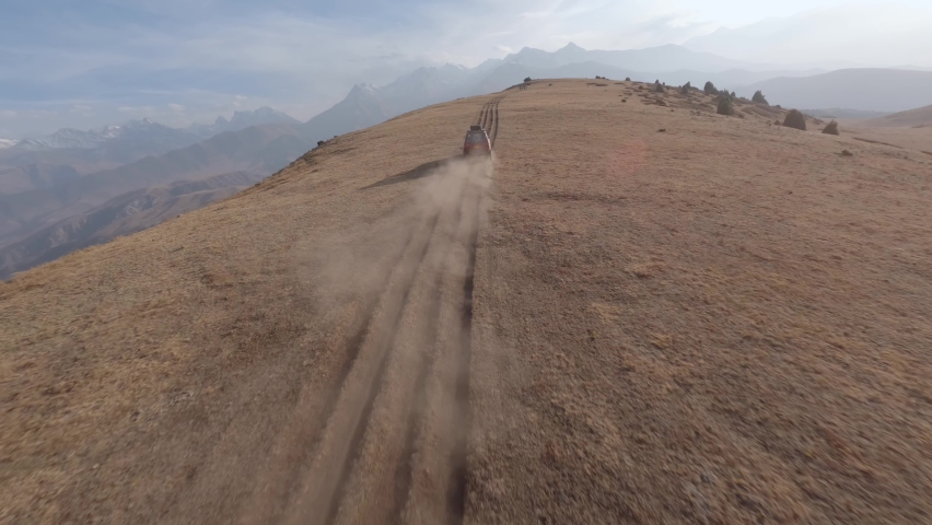 FPV sports drone follow shot SUV red automobile race rally offroad riding desert mountain adventure sand splash. Aerial view cinematic hilly valley dirt road transportation extreme tourism exploration