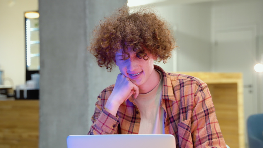 A young red -haired curly man with braces sits in a cafe smiles and looks at the camera. Young programmer in a cafe working on a laptop