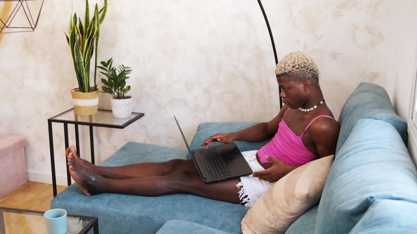 Serious African American person in feminine clothes working and typing on netbook keyboard while sitting on couch in sunlit living room at home