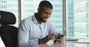 Happy millennial Black business man reading, typing text message on mobile phone, using app on Internet, chatting online, looking away, thinking, smiling, sitting at office work table