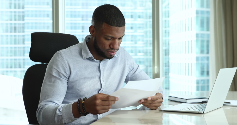 Serious focused African employee man holding document, receiving letter, reading legal paper, reviewing project report, sitting at laptop in office with panoramic window. Paperwork, business concept Royalty-Free Stock Footage #1096987841