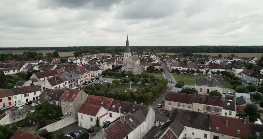 Aerial view of a church with bell tower in the heart of a small French village with a white house and a brown roof, a cloudy sky and a forest on the horizon. Royalty-Free Stock Footage #1096991327