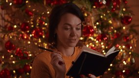 happy woman holding pen writing on the notebook. new year resolution or making notes in happiness journal on the background christmas tree with lights blinking and glowing - Powered by Shutterstock - Get 15% off with code: PIKWIZARD15