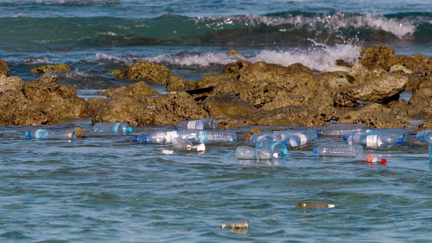 Empty plastic bottles and other rubbish float in sea between the stones near the seashore. Ecology of the earth, seas and oceans is in danger due to the large amount of garbage of human civilization