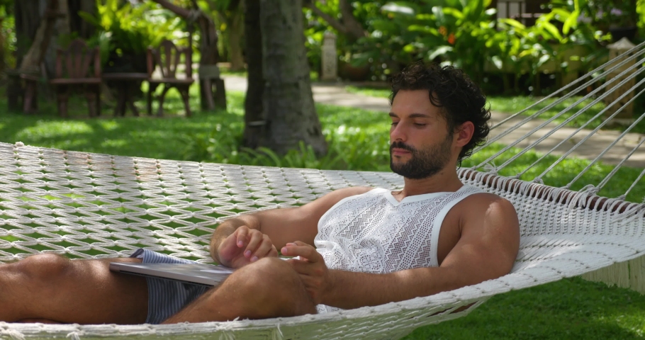 Charming guy, developer programmer, copywriter turning on a laptop PC while chilling in a beach hammock handing between two palm trees, telecommuting from anywhere in the world. Freelance occupation