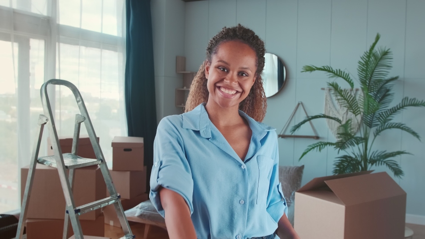 Young smiling African American woman showing off keys with keychain in shape of house showing off moving into spacious new apartment stands in living room with boxes and ladder. Realtor, homeowner