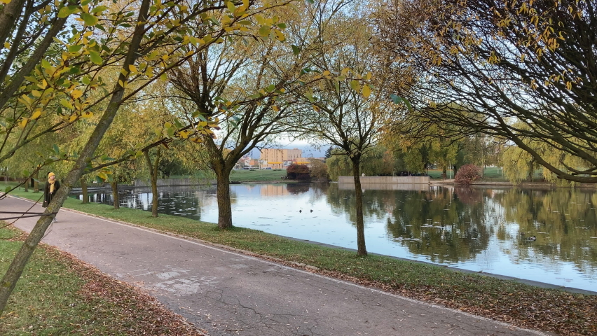 A young woman slowly walks through the park enjoying the nice autumn weather. Walk along the pond. In the water, a yellow-karsan leaf of trees is reflected