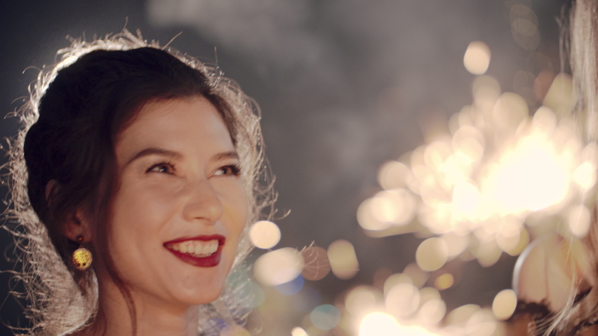 Beautiful woman holding christmas sparklers in hands. Friends celebrating by waving fireworks, enjoying party on New Years or Christmas Eve night. Happy group of people enjoying party in Xmas evening. | Shutterstock HD Video #1097020753