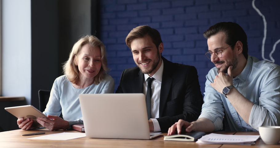 Three young and older colleagues talking sit at desk with laptop discuss new corporate software looking interested working on project, learn new business app, take part in teamwork, using modern tech Royalty-Free Stock Footage #1097021919