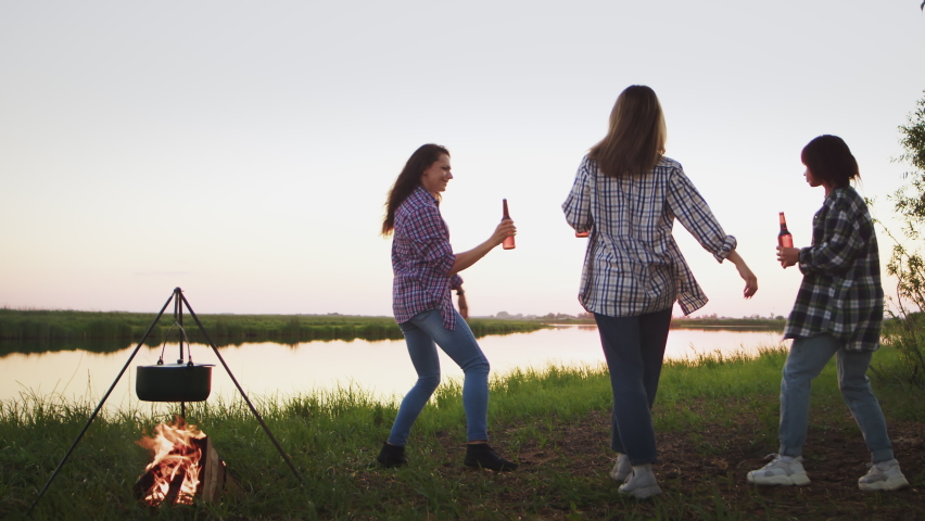 Happy ladies in checkered shirts dance on grassy river bank with bottles of beer at sunset twilight. Young women have fun on country picnic near burning campfire heating kettle in evening dusk