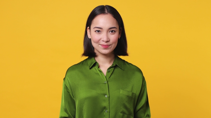 Beautiful charming happy fun young woman of Asian ethnicity 20s she wear green shirt posing looking camera smiling with sincere emotion isolated on plain yellow color wall background studio portrait