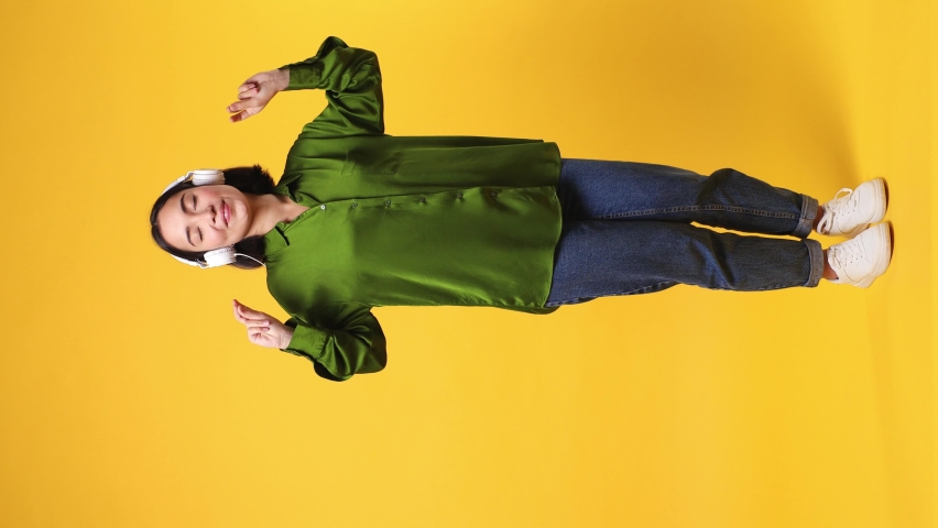 Full body vivid young woman of Asian ethnicity she wears green shirt listen mp3 music in headphones dance sing song have fun enjoy relax isolated on plain yellow color wall background studio portrait Royalty-Free Stock Footage #1097032039