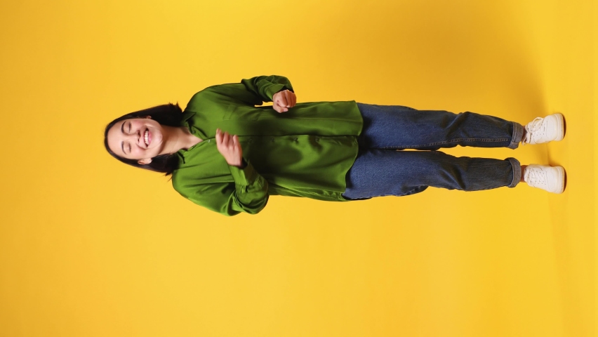 Full body fancy vivid happy bright young woman of Asian ethnicity she wear green shirt dance rising have fun enjoy relax celebrate play fluttering hair isolated on plain yellow color wall background