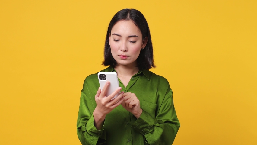 Sad upset young woman of Asian ethnicity 20s she wear green shirt hold use mobile cell phone swear read bad fake news unexpected rumor has some problem isolated on plain yellow color wall background Royalty-Free Stock Footage #1097032079