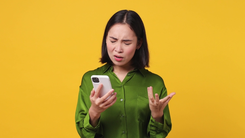Sad upset young woman of Asian ethnicity 20s she wear green shirt hold use mobile cell phone swear read bad fake news unexpected rumor has some problem isolated on plain yellow color wall background | Shutterstock HD Video #1097032079