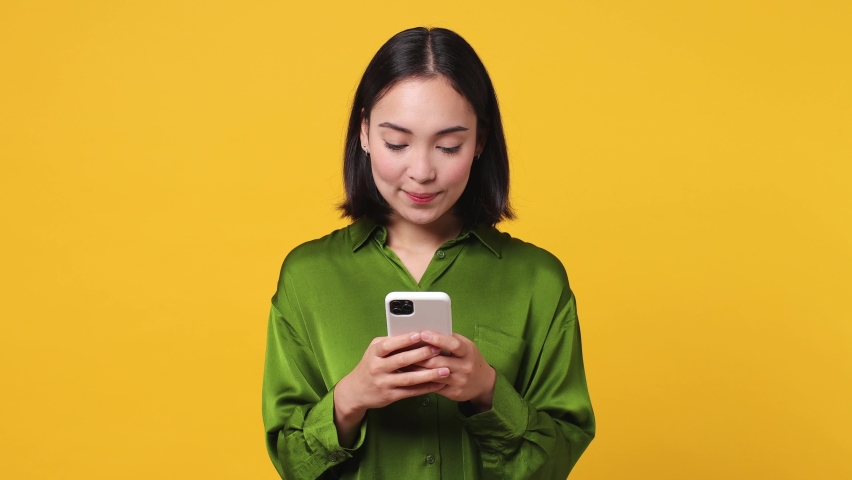 Happy smart joyful young woman of Asian ethnicity she wears green shirt hold use pointing finger on mobile cell phone just found out great big win news isolated on plain yellow color wall background