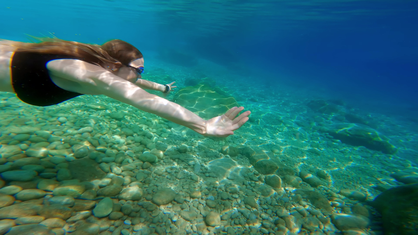 slender young woman in black bikini swims underwater near seabed. girl in black swimsuit swims forward, top view from under water. Journey travel to distant warm tropical countries.