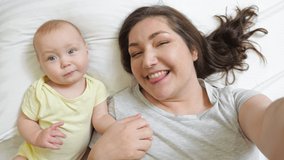POV of cheerful mom entertaining newborn baby by recording funny video and grimacing. Happy woman and daughter enjoy time together lying in bed closeup