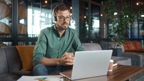 Elegant successful man working online using laptop wearing headphones video calling business meeting. Young handsome bearded male in glasses finishing work closing computer smiling. Video call concept