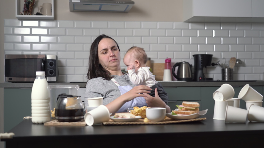 Tired mother trying to eat and drinking coffee while holding crying child in hands, woman after sleepless night because of baby in the messy kitchen | Shutterstock HD Video #1097052455