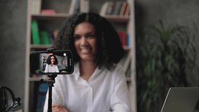 Blur background of pretty multinational woman with dark hair sitting at desk and filming video blog. Focus on modern digital phone fixed on tripod.