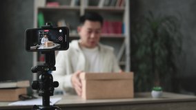 Focus on smartphone screen, happy asian male blogger unpacking gift boxes presents from companies and doing live streaming. Attractive man using smart phone camera for creating content.