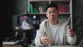 Smiling asian man in casual clothes sitting at desk with books, notes and laptop recording video blog and showing thumb up. Female tutor working at home during distance learning.