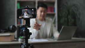 Focus on smartphone screen, pleasant young man writing report on laptop while recording video on his smart phone. Asian male freelancer working remotely while staying at home.