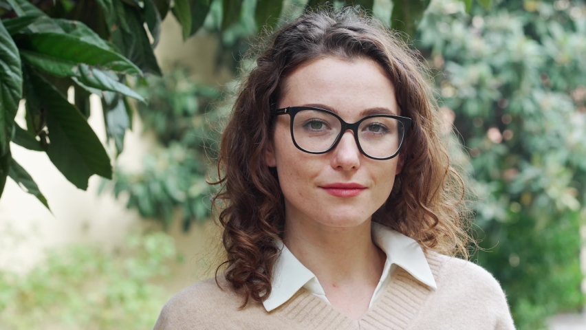 Young pretty confident professional business woman, proud businesswoman, beautiful curly lady wearing glasses standing outdoor on street, looking at camera, front face headshot portrait.