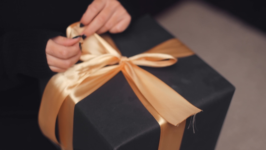 Close-up of a woman's hands tying a bow on a gift box in black and gold colors. Preparing for the Christmas holidays, giving gifts
