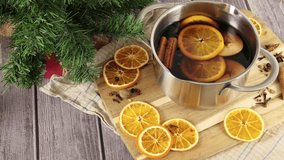Putting glass with mulled wine on wooden table decorated with Christmas tree, pot with hot red wine drink with cinnamon, apple and orange fruit and spice, festive alcohol, slow motion 4k video footage