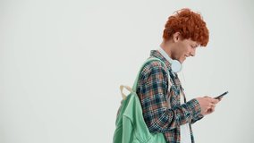 Cheerful red haired man with backpack typing on mobile and looking at the camera in the white studio - Powered by Shutterstock - Get 15% off with code: PIKWIZARD15
