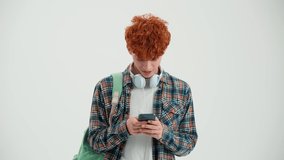 Laughing red haired man with backpack typing on phone and looking at the camera in the white studio - Powered by Shutterstock - Get 15% off with code: PIKWIZARD15