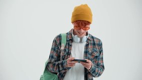 Positive red haired man with backpack playing game on mobile in the white studio - Powered by Shutterstock - Get 15% off with code: PIKWIZARD15