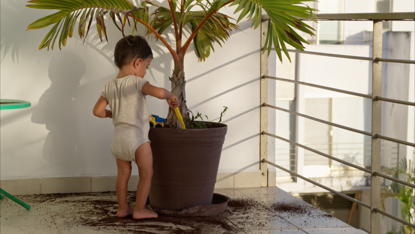 Little latin baby boy scooping dirt from a plant pot and dropping it to the ground with a green plastic kitchen spoon. Mischief Royalty-Free Stock Footage #1097096955