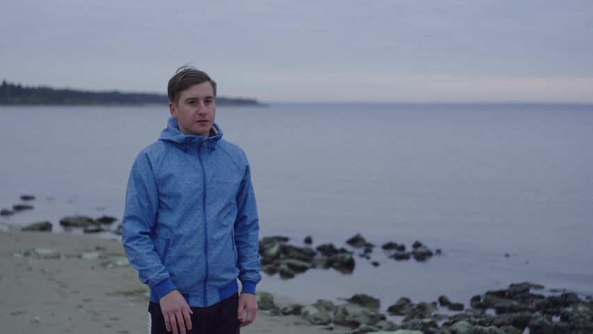 Portrait, morning run of a man in blue clothes near a reservoir, in cloudy weather. Sportsman jogging near the river. Sports near the lake. Running along the coastline of the sea, autumn. 4k