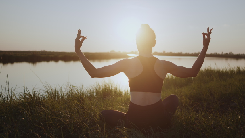 Young woman in sportswear spreads hands putting in prayer gesture at bright sunset light in evening. Lady enjoys practicing yoga exercise on river bank sitting in Lotus pose at twilight backside view | Shutterstock HD Video #1097104199