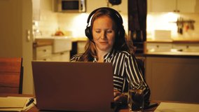 Female wearing headphones looking at laptop screen, having class online in the evening in cozy kitchen, Young woman student study learn online with computer, wearing earphones, talking to teacher