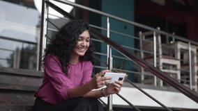 Beautiful Indian girl holding smart phone watching funny videos ,movie or social media content while sitting in stairs. Pretty woman looking at mobile screen and watching something funny video.