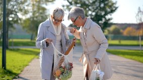 Two senior female friends are holding grocery bags and walking through park