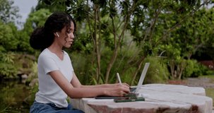 Young African American female teenager sitting in an online class on a laptop and taking notes on a tablet in the park. Working outside with high-speed Wi-Fi internet communication technology