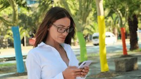 smiling young Latin beautiful woman sitting outside in city looking at mobile phone. brunette excited overjoyed latin happy cute woman 30s in White shirt hold mobile cell phone chat typing sms
