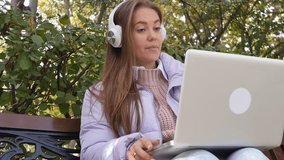 Portrait of a charming young woman with white headphones and a laptop talking on a video call while sitting on a park bench and holding a computer on her legs. Video call communication remotely